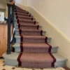 Red herringbone flatweave stair runner with a woven red border installed on a wooden winding staircase.