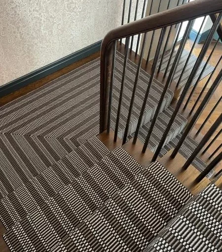 Tapis d'escalier à rayures noires, blanches et grises, installé sur des escaliers en bois et un palier.