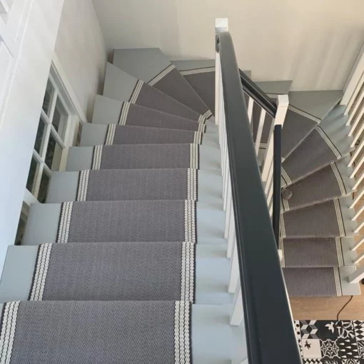 Tapis d’escalier gris et blanc avec bordure tissée, installé sur un escalier en bois en colimaçon.
