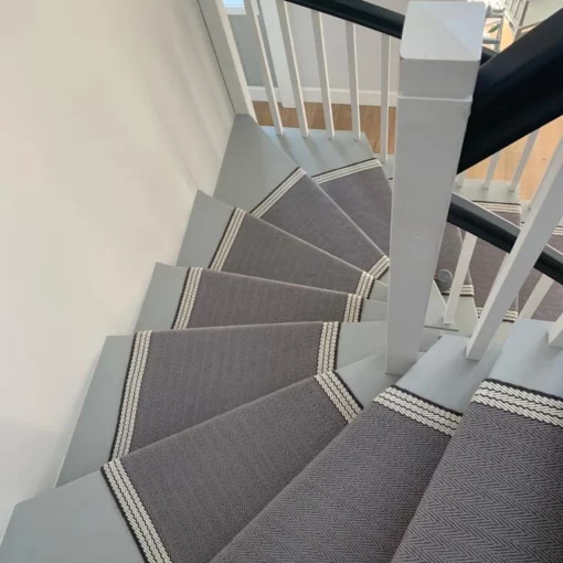 Tapis d’escalier gris et blanc avec bordure tissée, installé sur un escalier en bois en colimaçon.